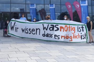 Sechs Aktivist*innen stehen vor dem Veranstaltungsort des Städtetages BW 2022, dem SNP Dome in Heidelberg. Sie halten ein Banner mit der Aufschrift "Wir wissen, was nötig ist und wir wissen, dass es möglich ist" hoch.